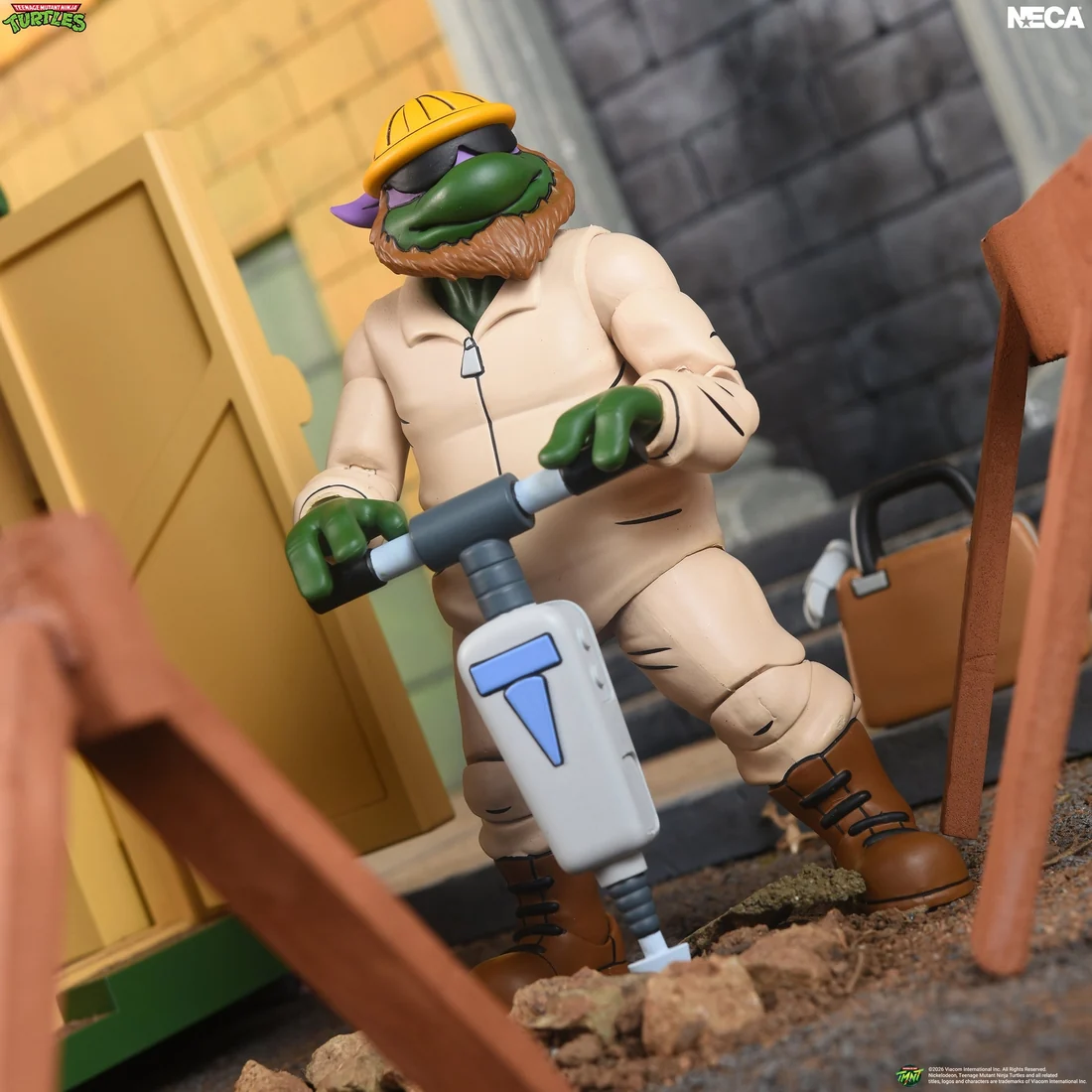 Construction Don in tan coveralls, yellow hard hat, sunglasses and a ginger beard, leaning on a jackhammer beside a sawhorse.